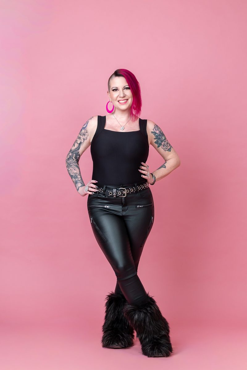 Beth Carter smiling, hands on hips, pink hair and all-black outfit against a pink background
